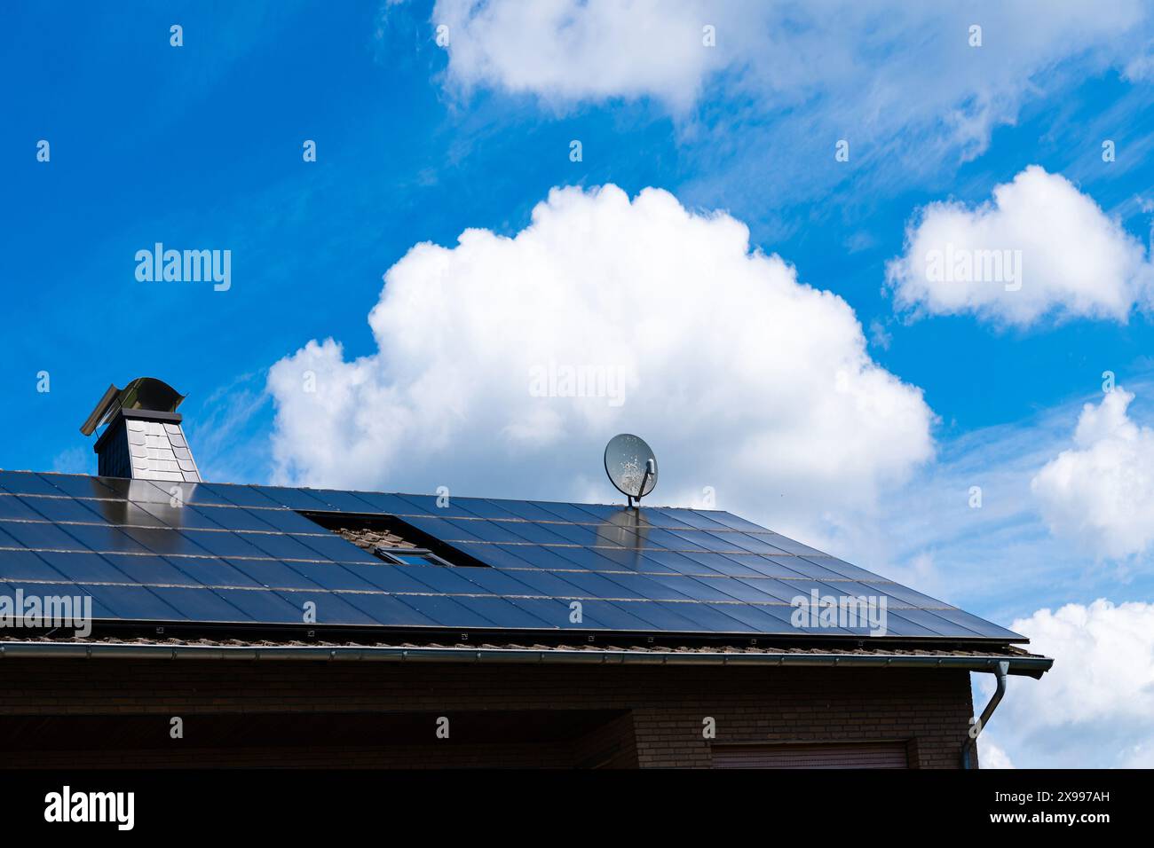 Das Dach des Gebäudes mit einem Kamin und einer Satellitenschüssel ist mit Solarpaneelen bedeckt. Blauer Himmel mit schönen weißen Wolken. Stockfoto