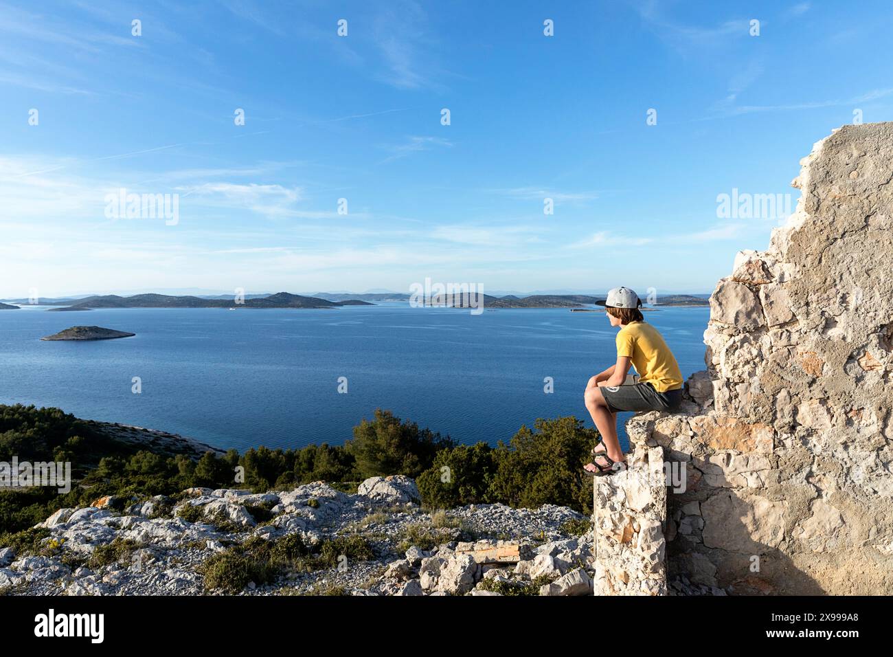 Junge, Tourist, der auf einer Ruine eines Militärgebäudes auf dem Hügel auf der Insel Žirje sitzt, einer kleinen Insel in der Mitte der Adria, Kroatien Stockfoto