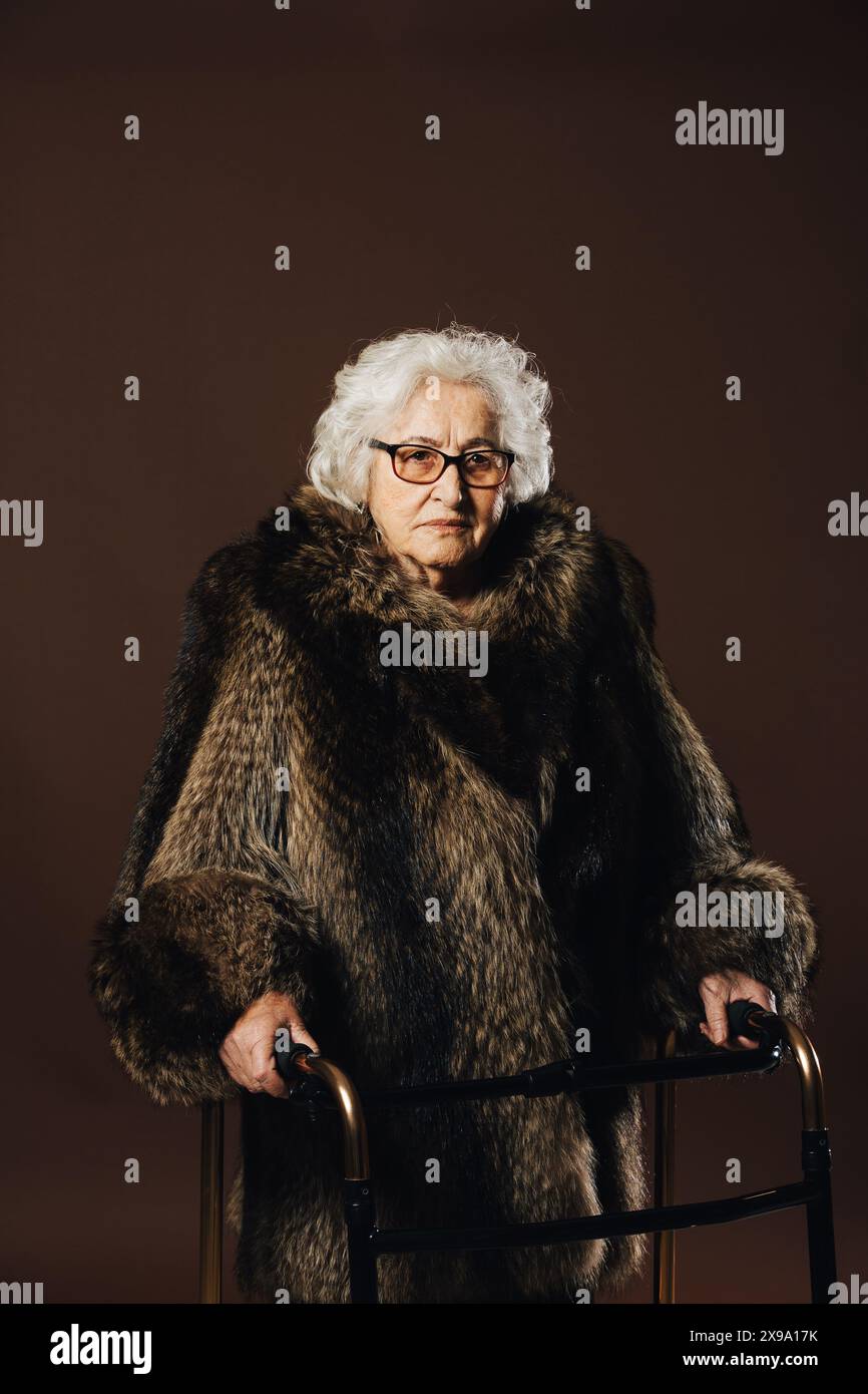 Eine ältere Frau mit lockigem weißem Haar und Brille steht und hält einen Geher in einem Pelzmantel vor einem schlichten dunklen Hintergrund. Stockfoto