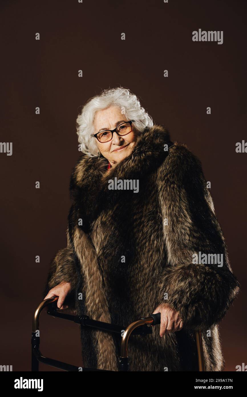 Eine ältere Frau mit lockigem weißem Haar und Brille steht und hält einen Geher in einem Pelzmantel vor einem schlichten dunklen Hintergrund. Stockfoto