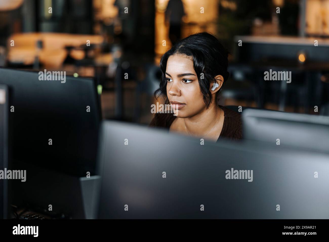 Junge Unternehmerin mit kabellosen in-Ear-Kopfhörern, die im Büro am Computer arbeiten Stockfoto