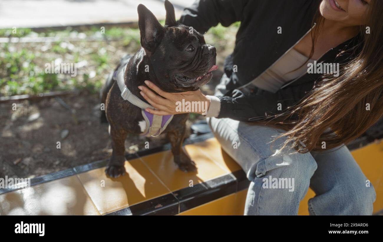 Eine junge hispanische Frau streichelt ihre französische Bulldogge draußen in einem Stadtpark und unterstreicht die Bindung zwischen Haustier und Besitzer in einer Stadt. Stockfoto