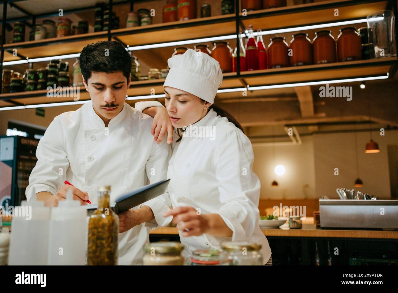 Weibliche Kochhand auf der Schulter eines männlichen Küchenchefs, der im Tagebuch schreibt, während er in der kommerziellen Küche steht Stockfoto