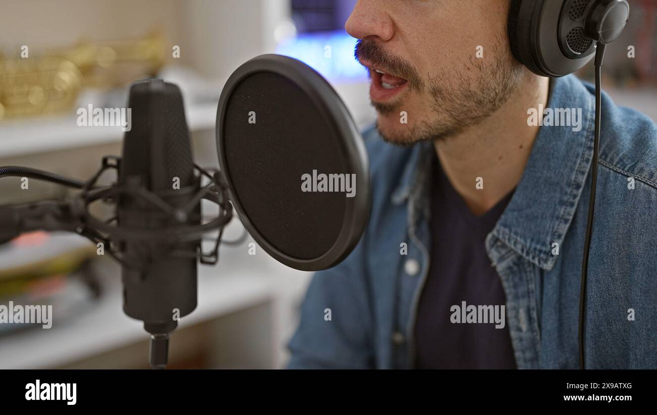 Hispanischer Mann mit Bart, der drinnen in einem Musikstudio singt und Mikrofon und Kopfhörer zeigt. Stockfoto