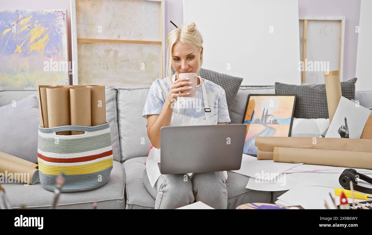 Lebendige, junge blonde Künstlerin schlürft bei einem ansprechenden Videoanruf in ihrem geschäftigen Studio Kaffee Stockfoto