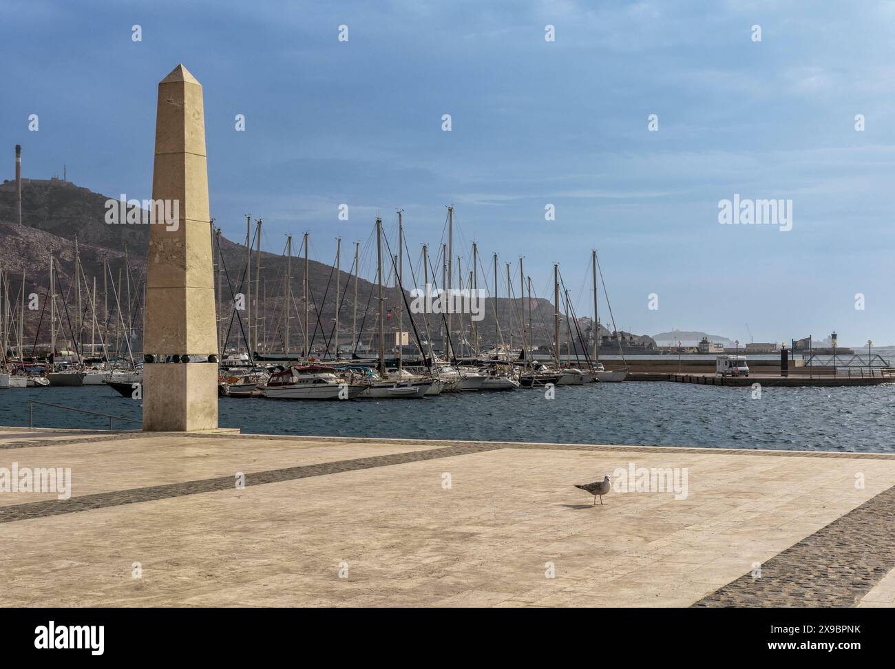 Foto einer Frau, die entlang der Esplanade des Hafens der Stadt Cartagena zwischen den beiden Säulen läuft, mit Booten, die im Hintergrund verankert sind, Murcia. Stockfoto