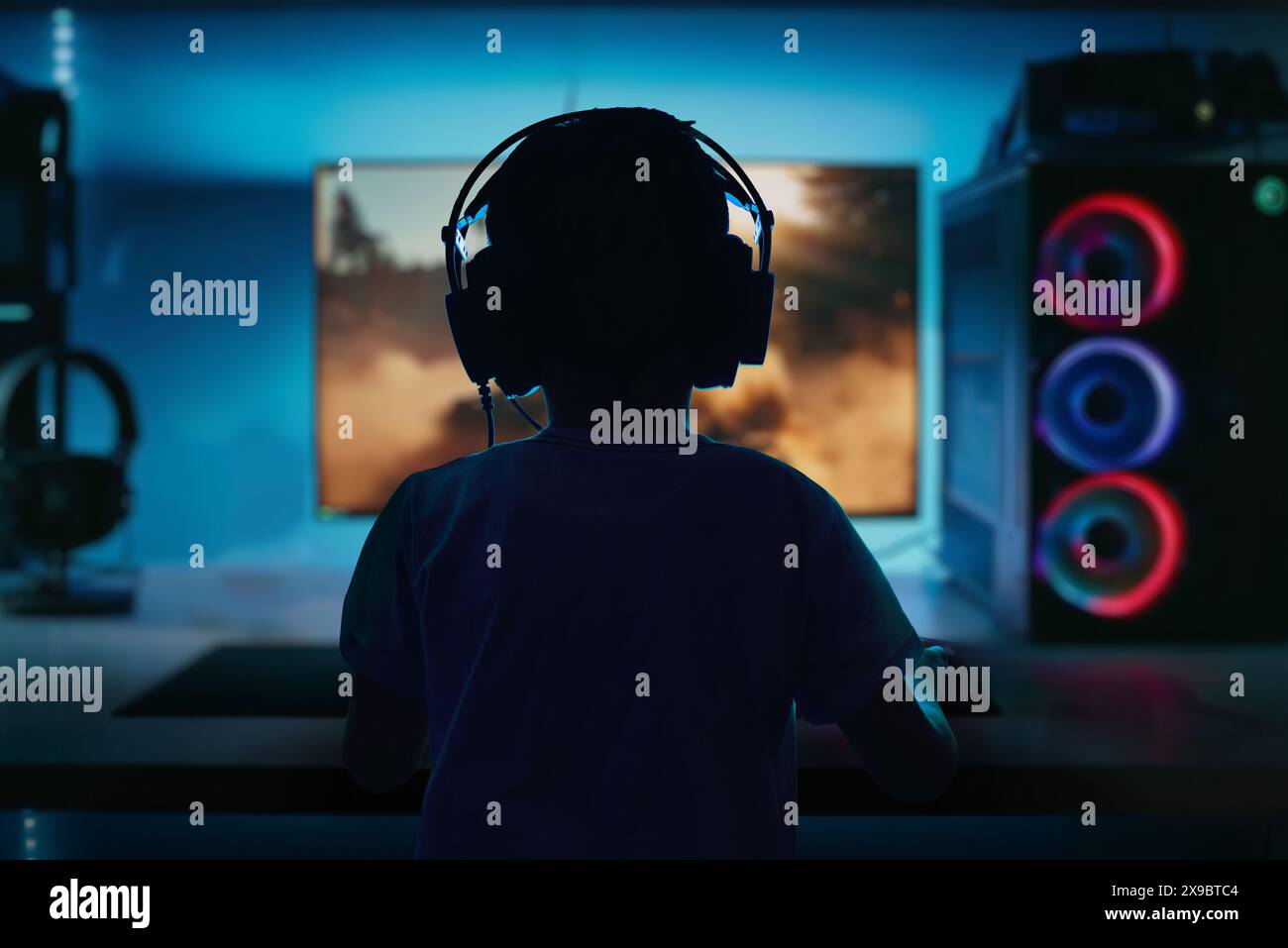 Schwaches, kontrastreiches Bild eines Jungen, der ein Computerspiel auf einem Gaming-PC spielt, von hinten betrachtet, mit einer dramatischen Auswahl an LED-Leuchten Stockfoto