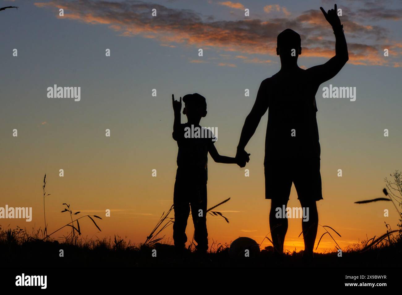 Silhouetten im Sonnenuntergang von Vater und Sohn, die Hände halten und mit der anderen Hand auf dem Feld ein Felssymbol machen. Stockfoto