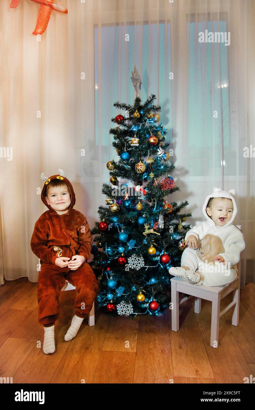 Zwei Kinder in Kostümen sitzen an einem geschmückten Weihnachtsbaum in einem gemütlichen Zimmer mit Holzfußboden Stockfoto