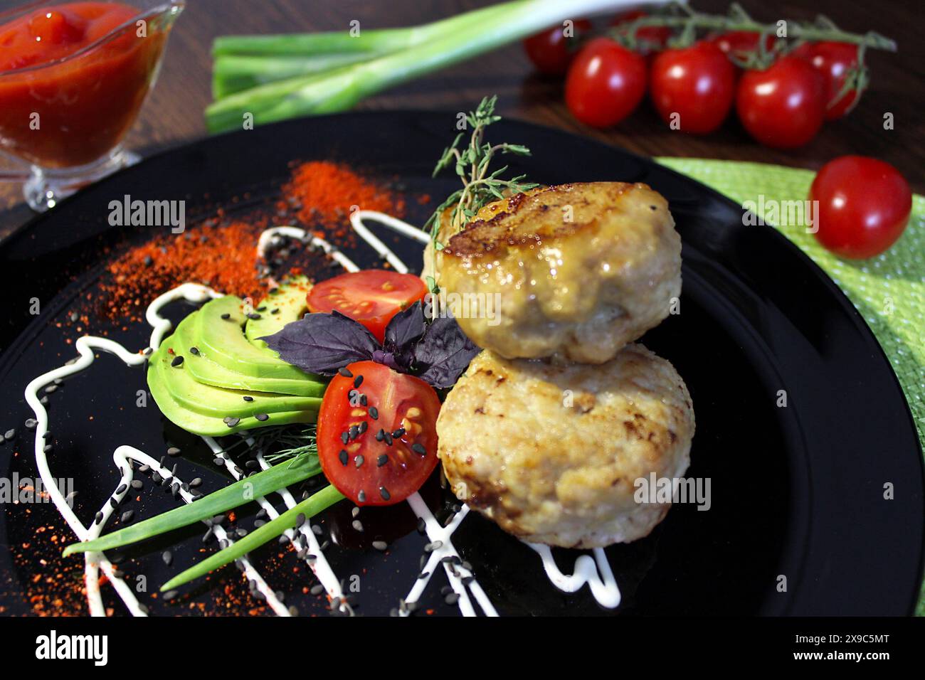 Gourmet-Gericht mit Fleischbällchen, serviert mit geschnittener Avocado, Kirschtomaten und Kräutern auf einem schwarzen Teller mit künstlerischem Sauce-Nieseln und rotem Pfefferpulver Stockfoto