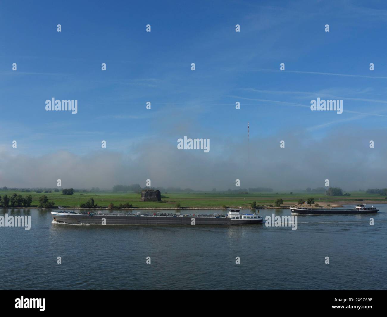 Mehrere Schiffe auf einem Fluss in einer grünen, naturbelassenen Landschaft unter blauem Himmel, Schiffe auf dem rhein am frühen Morgen, wesel, Deutschland Stockfoto