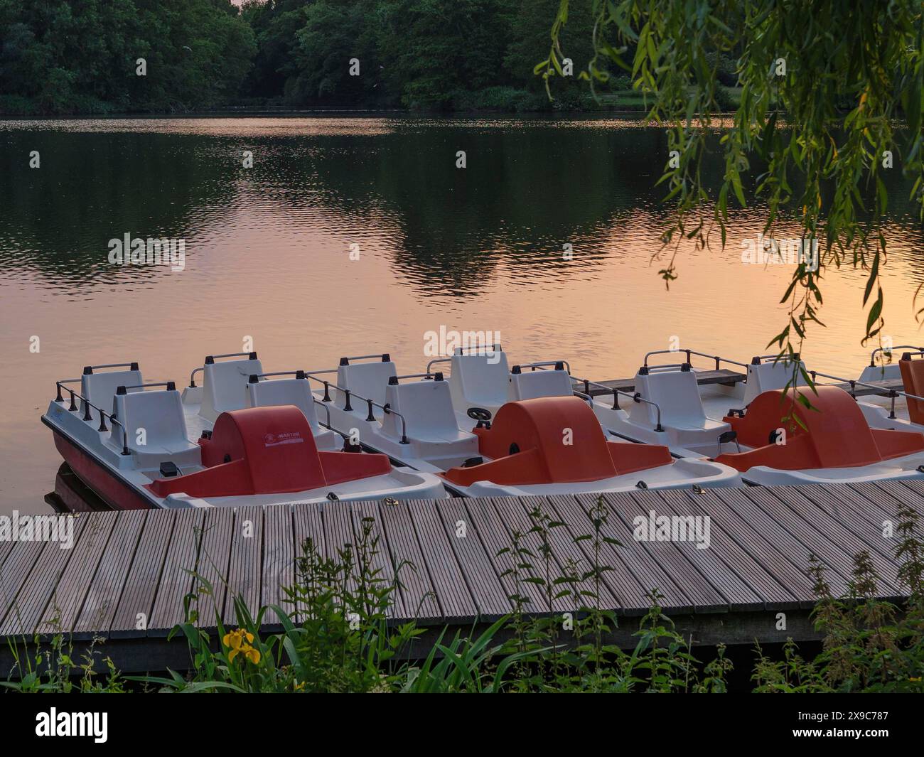 Eine Gruppe von Tretbooten ist an einem Steg verankert, der sich in einen See erstreckt. Der Himmel im Hintergrund zeigt die Farben des Sonnenuntergangs, Steg an einem kleinen Stockfoto