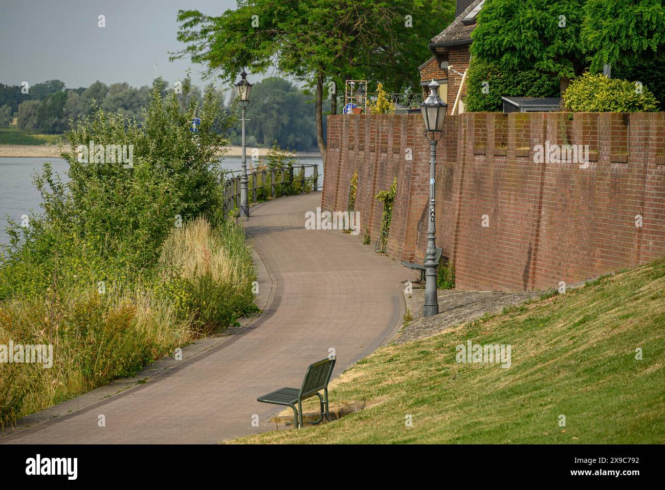 Kurviger Weg entlang einer Backsteinmauer mit Parkbänken und Lampen, gesäumt von grünen Bäumen und Häusern, die Promenade von Rees am Rhein mit grünen Bäumen Stockfoto