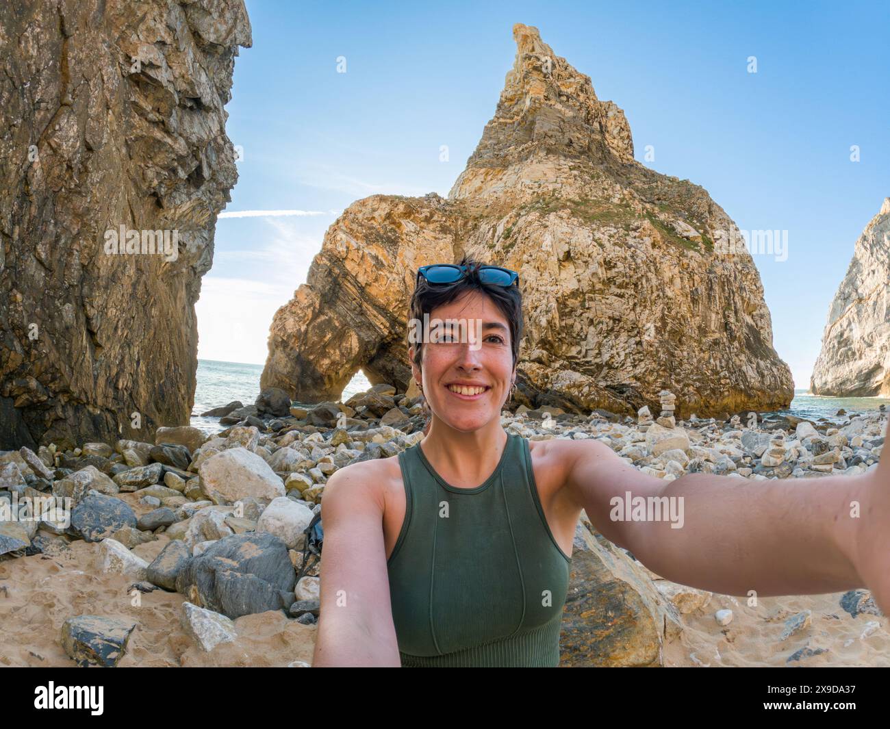 Frau am Strand macht ein Selfie bei Sonnenuntergang mit Bergen im Hintergrund, das Meer lächelnd und in die Kamera schauend Stockfoto