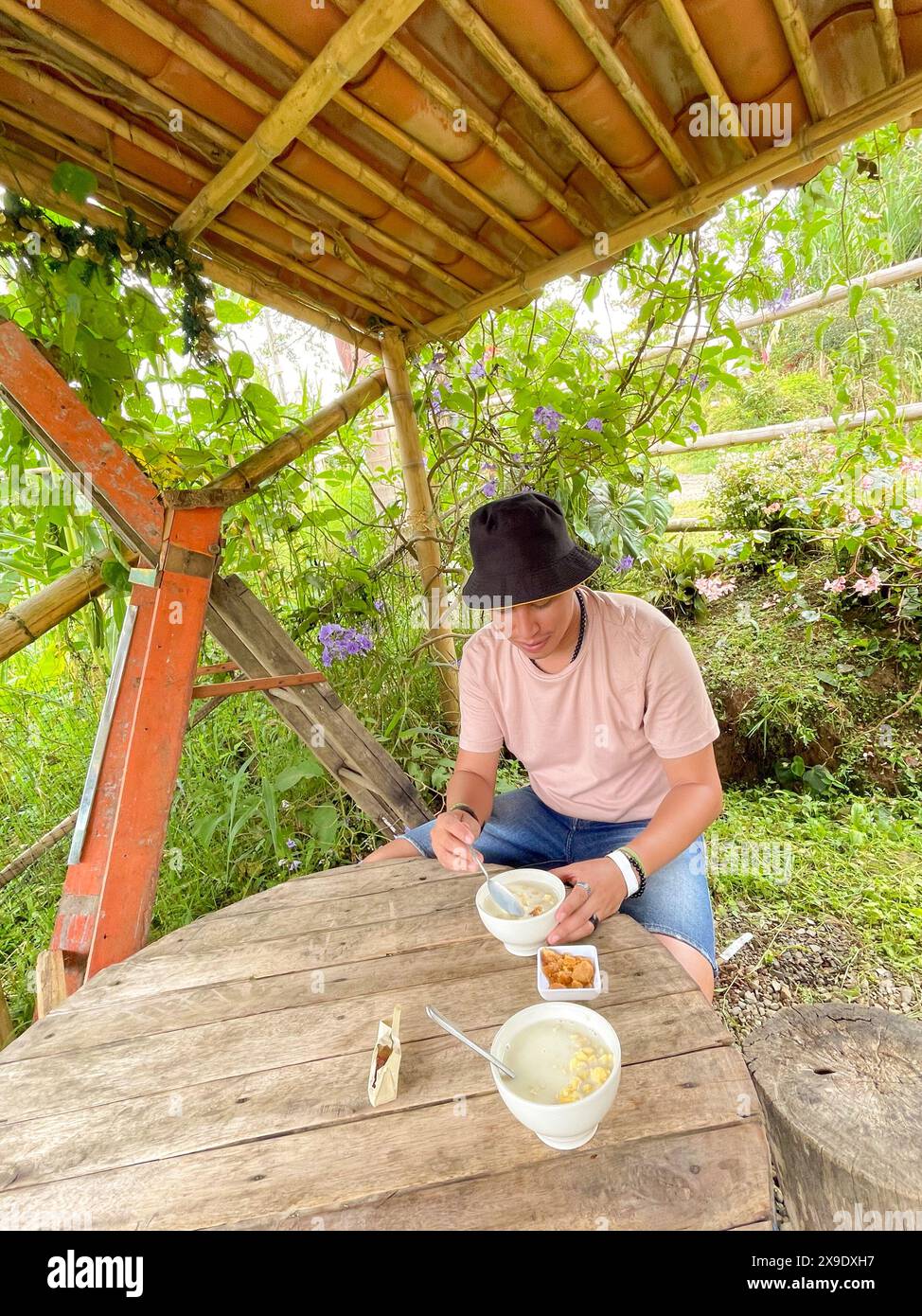 Lässig gekleideter Mann sitzt an einem rustikalen Holztisch unter einem Bambusunterstand und genießt eine Mahlzeit umgeben von üppigem Grün. Stockfoto