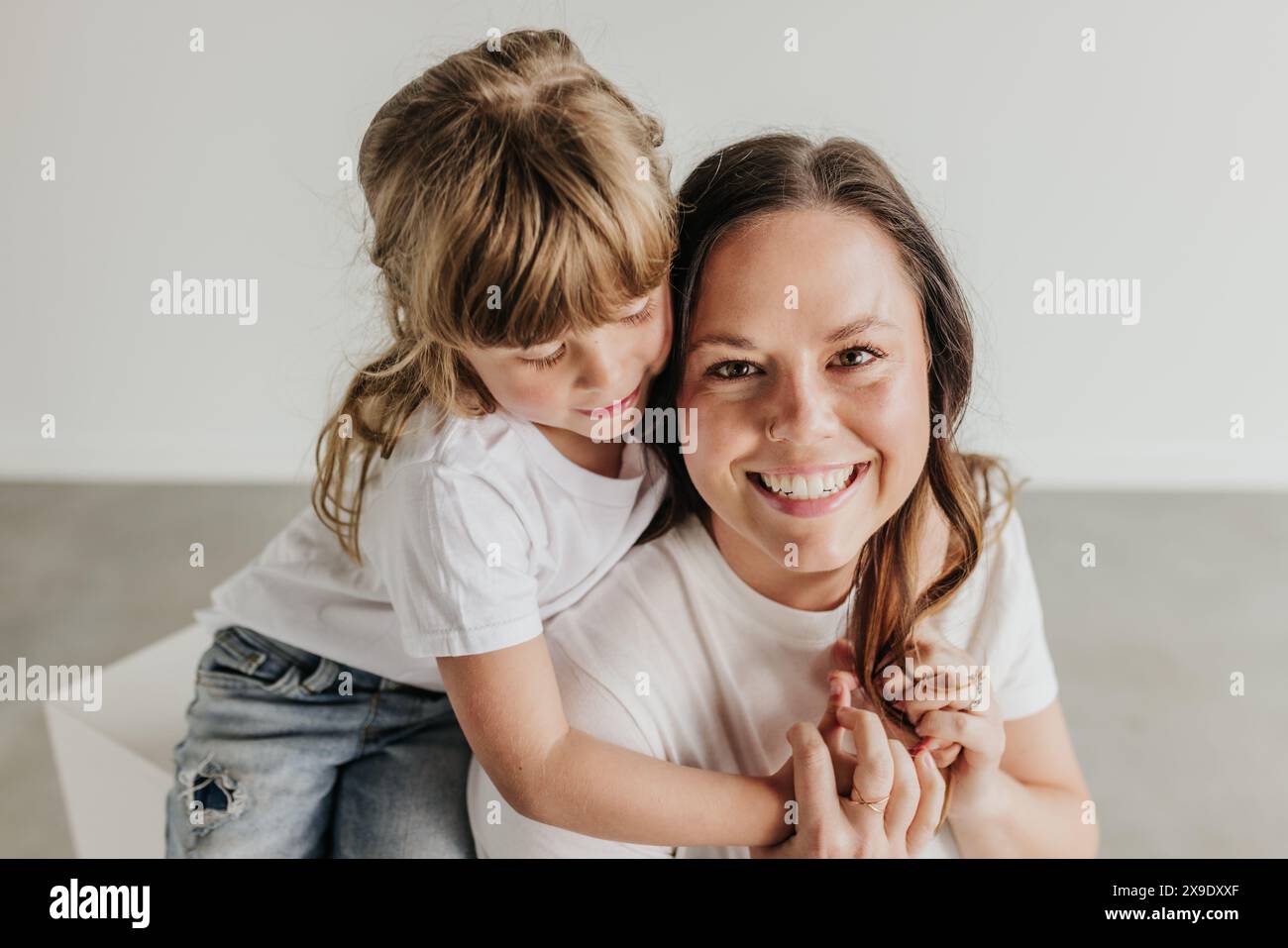 Die junge Tochter umarmt die lächelnde Mutter von hinten im Studio Stockfoto