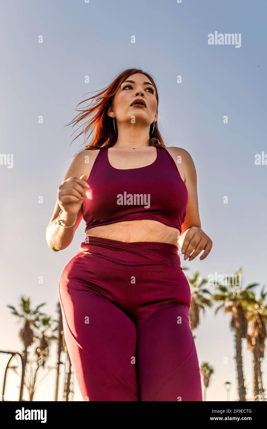 Frau in Übergröße, die im Freien in kastanienbrauner Sportbekleidung läuft, Entschlossenheit und Fitness-Motivation demonstriert, gesunder Lebensstil, heller und sonniger Tag, ac Stockfoto