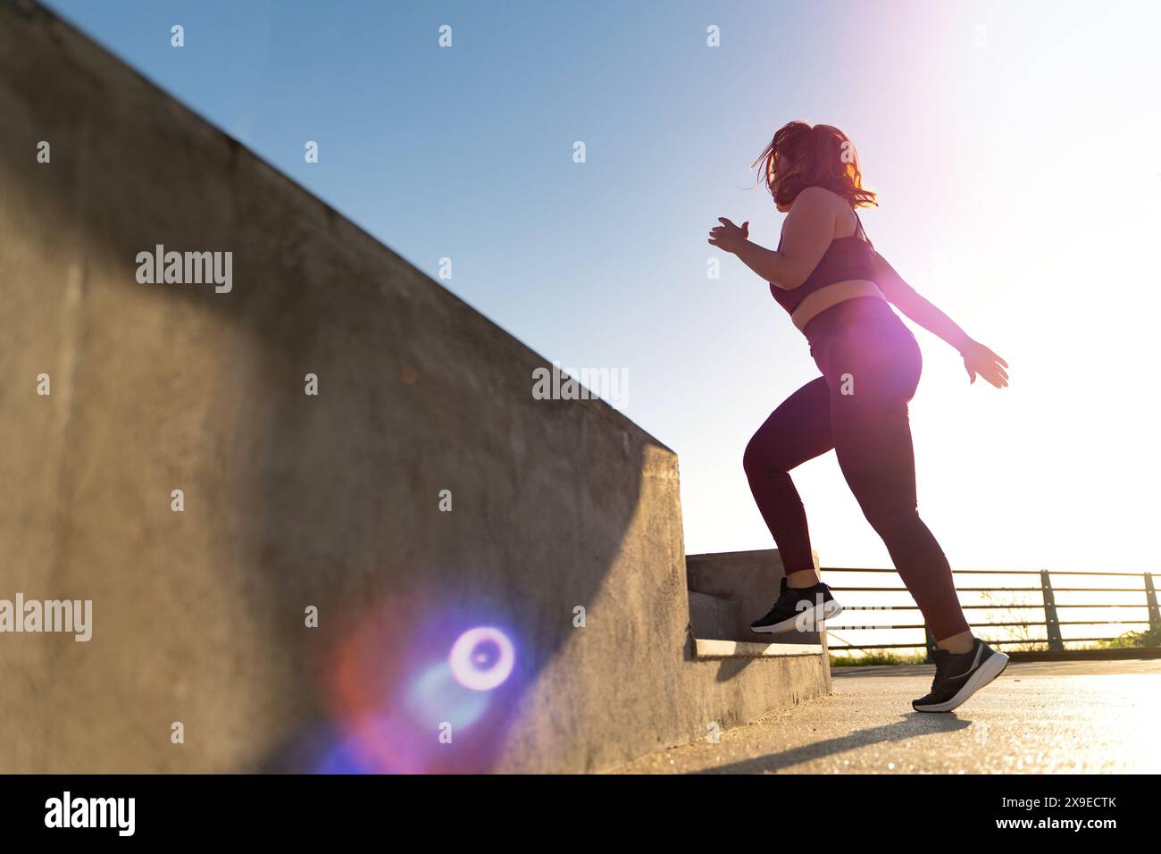 Plus-Size Frau, die während eines Outdoor-Workouts mit Linsenflare-Effekt Treppen hinauf läuft, - Entschlossenheit und Fitness-Motivation, gesunder Lebensstil, hell Stockfoto