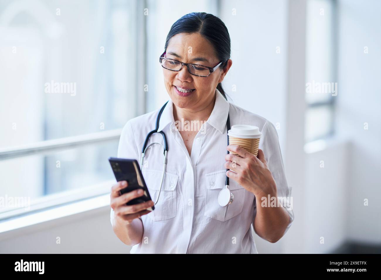 Lächelnder Arzt macht eine Pause mit Kaffee und Smartphone im Krankenhaus Stockfoto