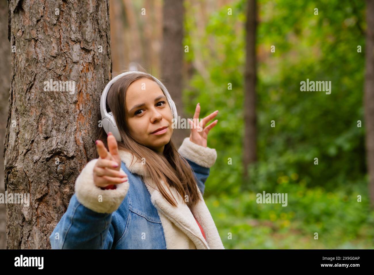 Eine junge Frau lehnt sich an einen Baum, trägt Kopfhörer und genießt Musik im Wald an einem sonnigen Nachmittag Stockfoto