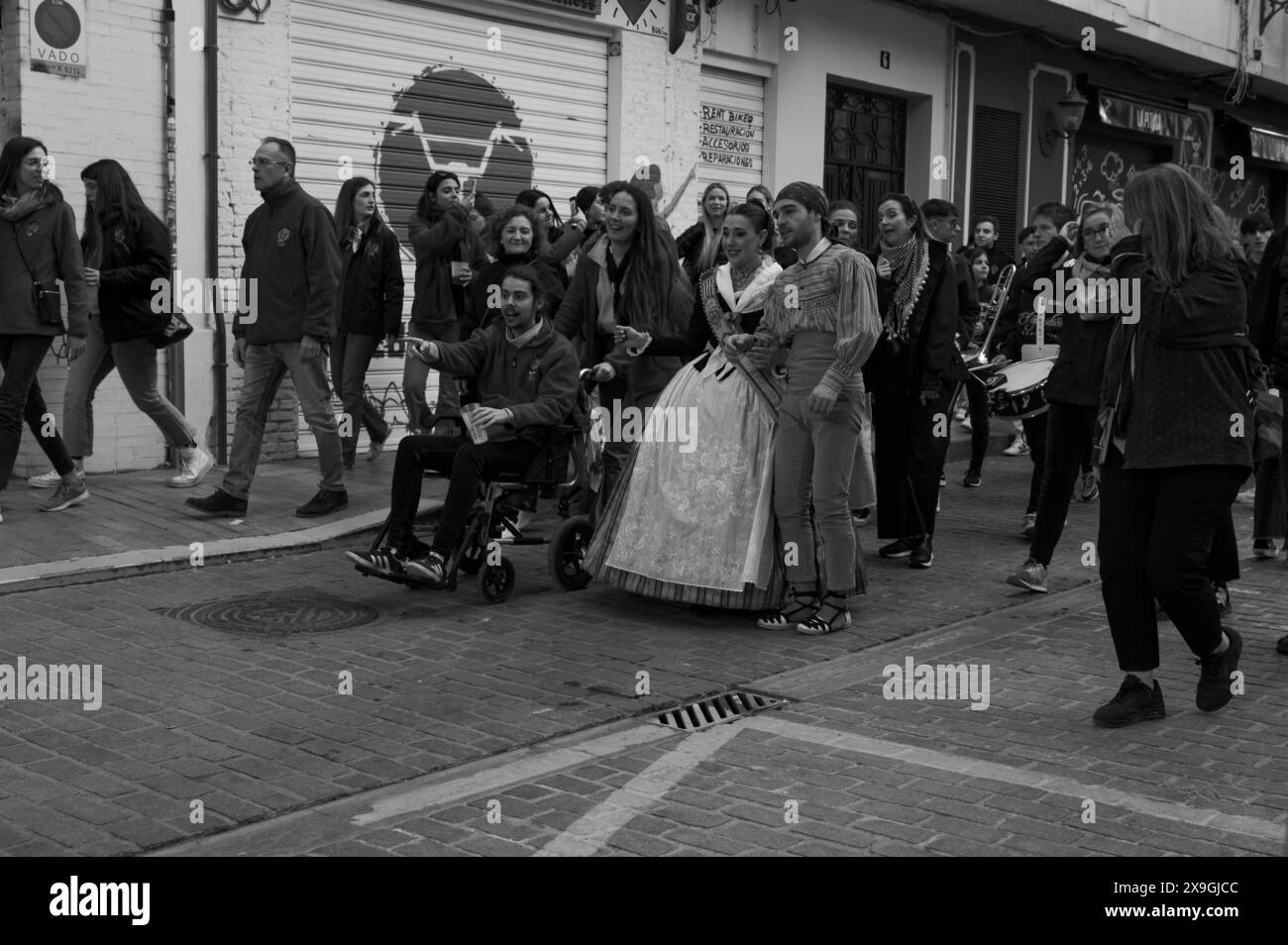 Festliche Prozession in Valencia mit Menschen in traditioneller Fallera- und Fallero-Kleidung, begleitet von Einheimischen, darunter ein Mann im Rollstuhl Stockfoto