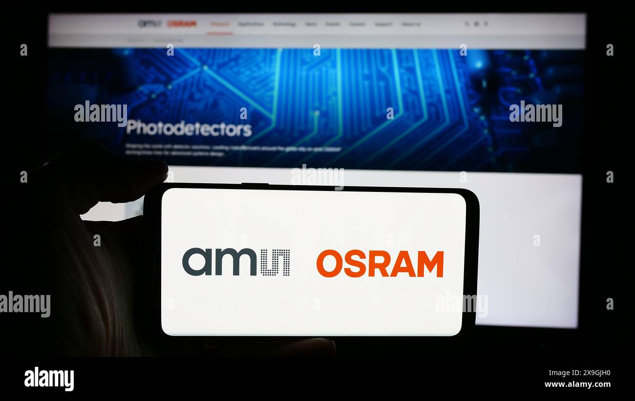 Person, die ein Mobiltelefon mit dem Logo des österreichischen Elektronikunternehmens ams OSRAM AG vor der Business-Website hält. Schwerpunkt auf dem Display des Telefons. Stockfoto