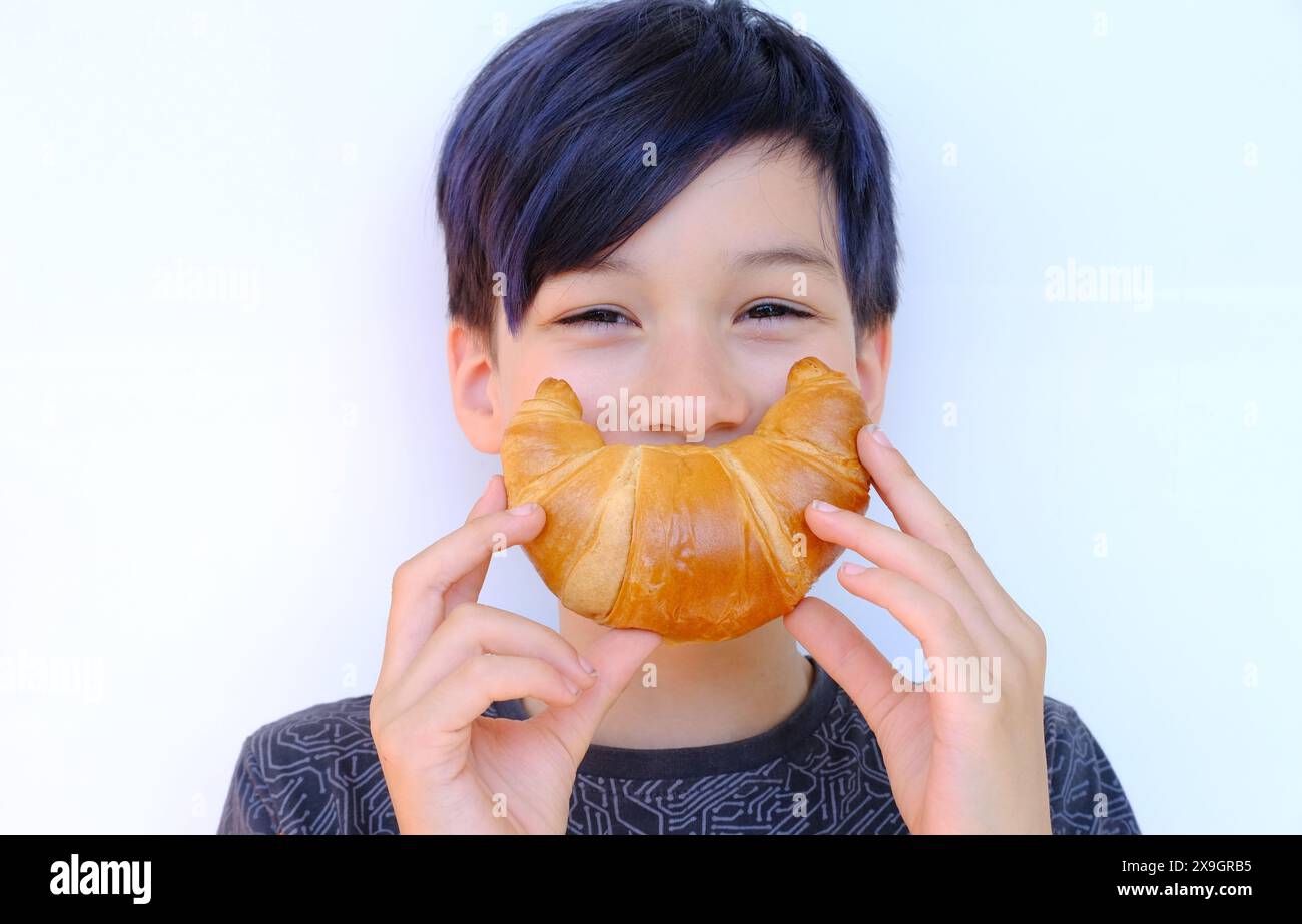 Nahaufnahme des Gesichts des Kindes, Junge von 10 Jahren hält rosa gebackenes Croissant mit Händen nahe dem Mund, zeigt fröhliches Lächeln, Konzept köstliches Frühstück, h Stockfoto