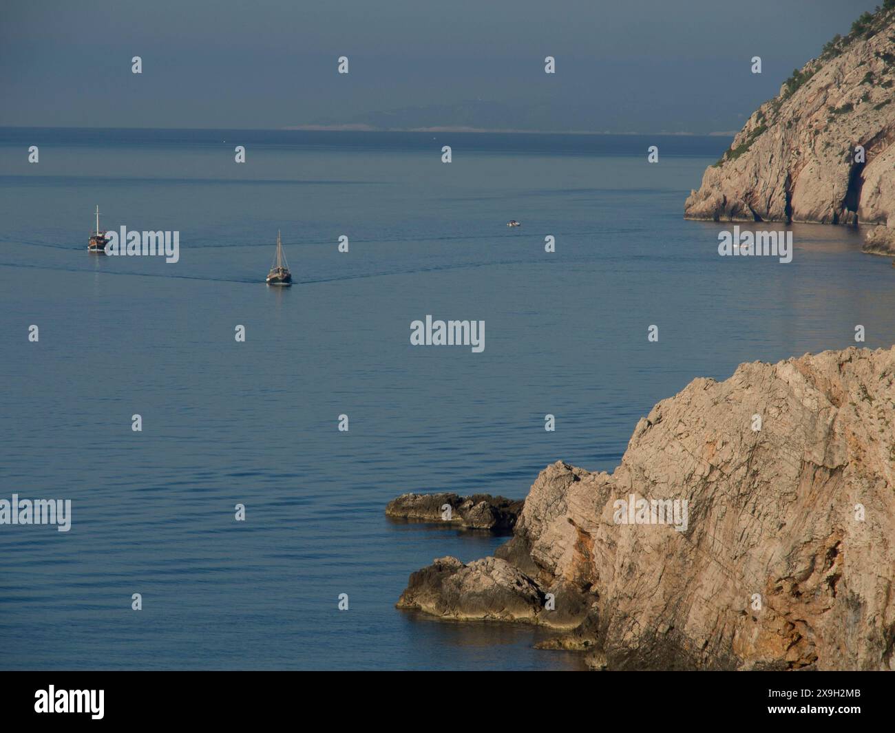 Zwei Boote segeln auf dem ruhigen Meer entlang einer felsigen Küste, der Altstadt von Dubrovnik mit historischen Häusern, Kirchen, roten Dächern und Festungsmauern Stockfoto