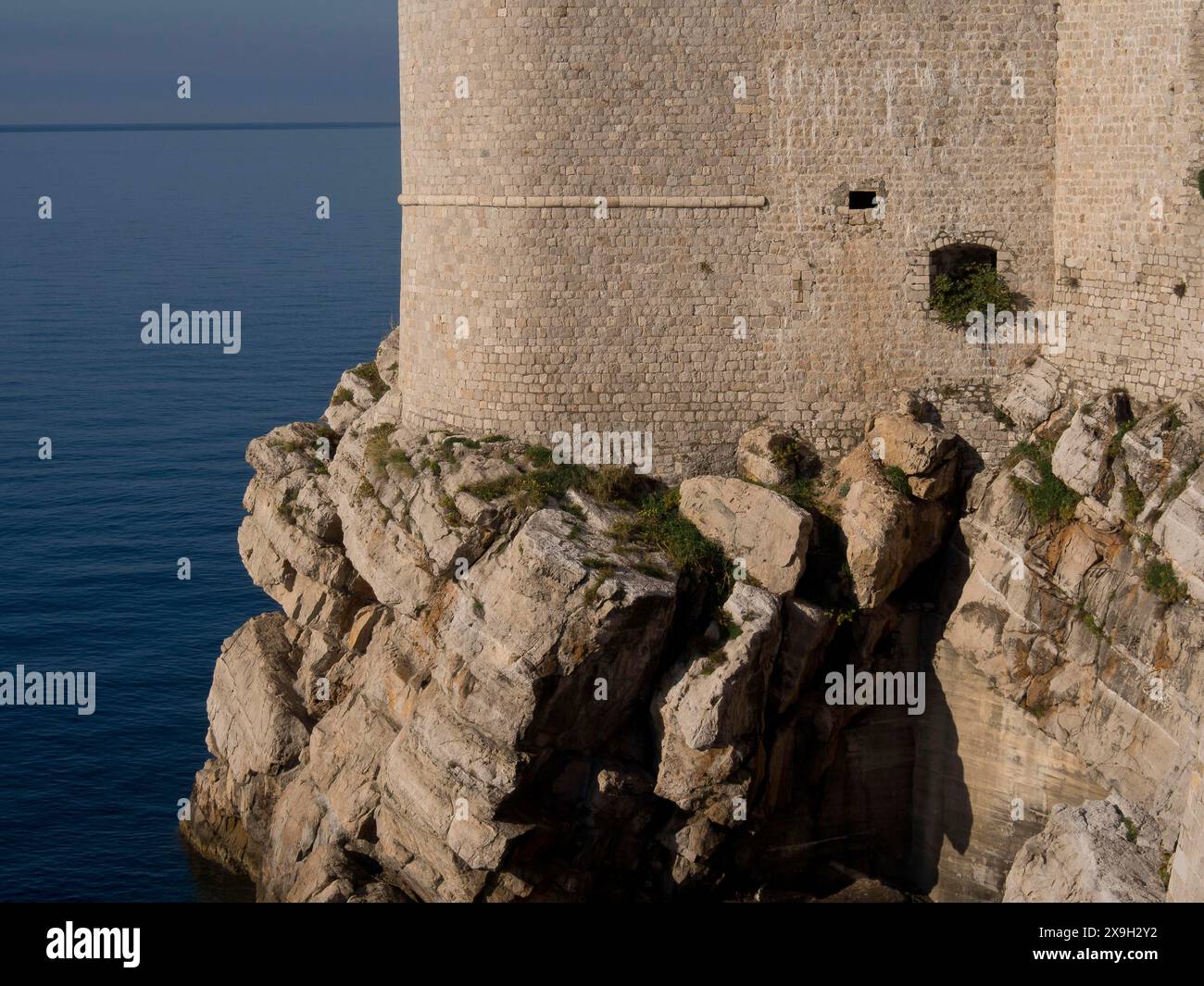 Die robuste Küstenmauer erhebt sich vom felsigen Ufer und überblickt das Meer, die Altstadt von Dubrovnik mit historischen Häusern, Kirchen, roten Dächern und Stockfoto