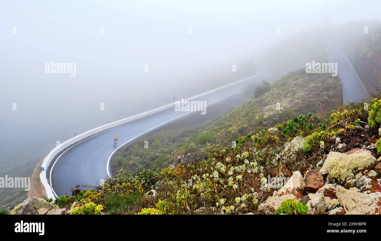 Nebelige Bergstraße mit einem einsamen Radfahrer, der sich durch üppige grüne Landschaft schlängelt Stockfoto