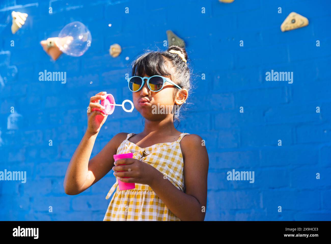 Ein birassisches Mädchen, das eine Sonnenbrille und ein Sommerkleid trägt, bläst gern draußen Blasen, mit Kopierraum Stockfoto