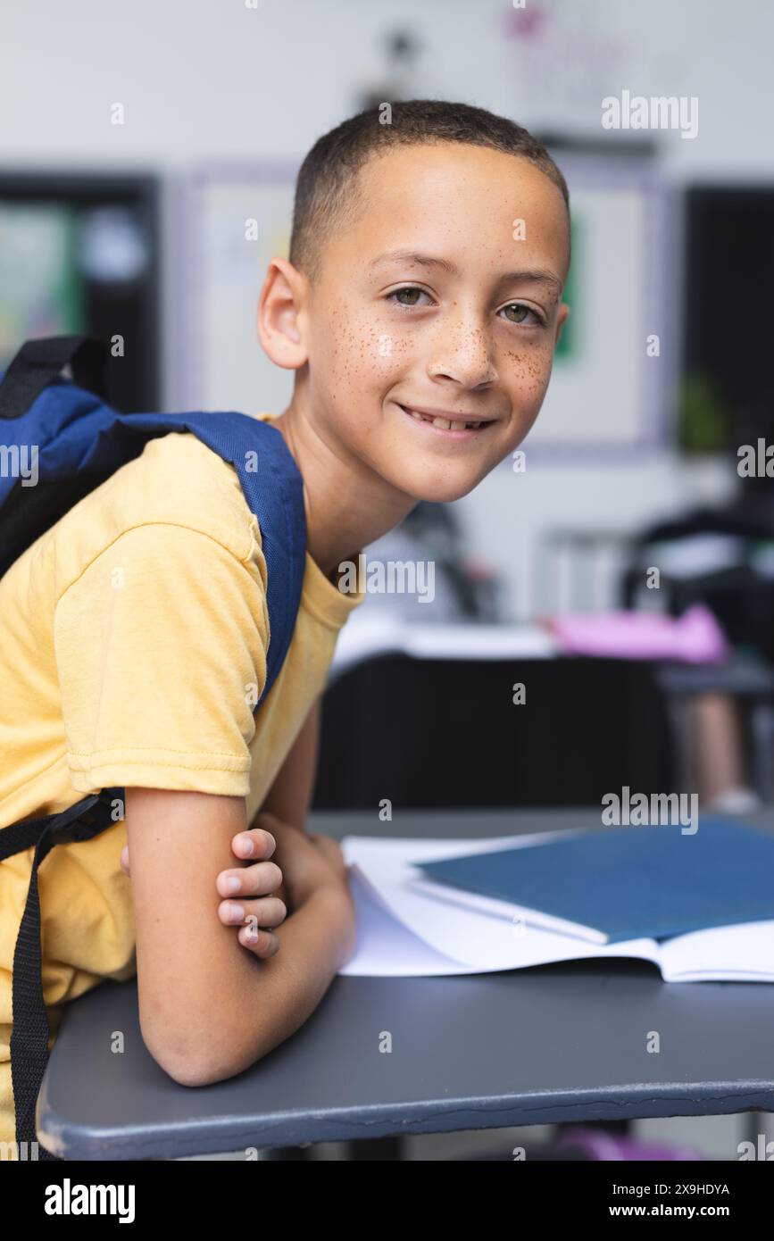 Ein birassischer Junge mit Rucksack sitzt an einem Schreibtisch in einem Klassenzimmer und lächelt Stockfoto