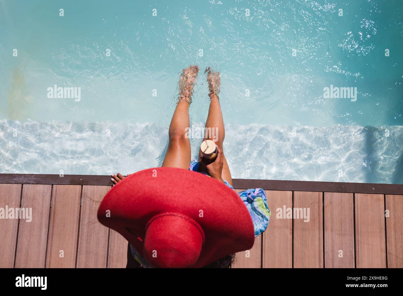 Eine zweirassige junge Frau, die einen roten Hut trägt und draußen im Poolwasser die Füße hängt Stockfoto