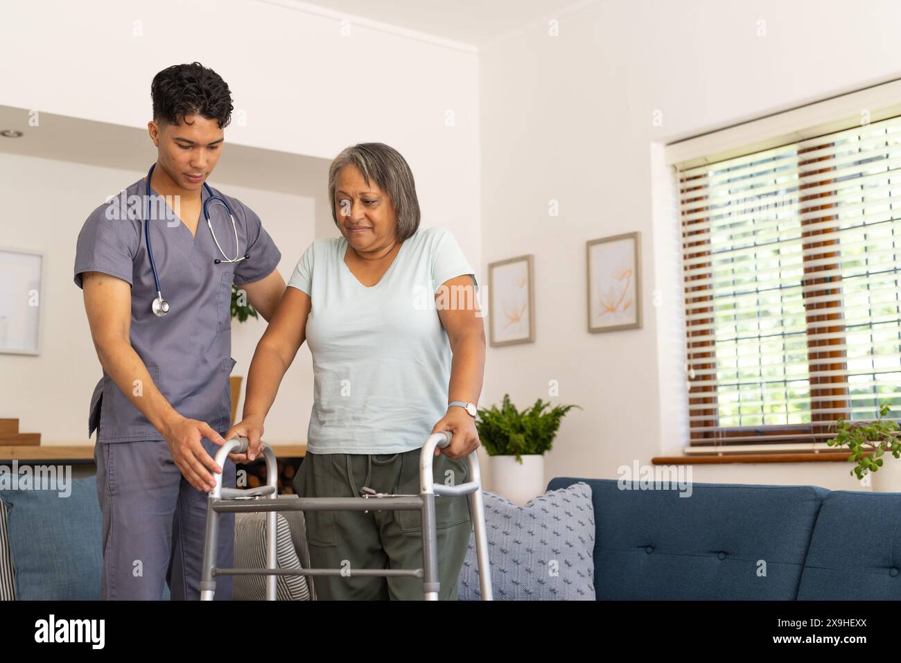 Zu Hause, junge birassische männliche Krankenschwester, die älteren weiblichen Patienten hilft. Sie sind in hellem, gemütlichem Wohnzimmer mit blauem Hintergrund, Sofa und großer Fensterscheibe Stockfoto