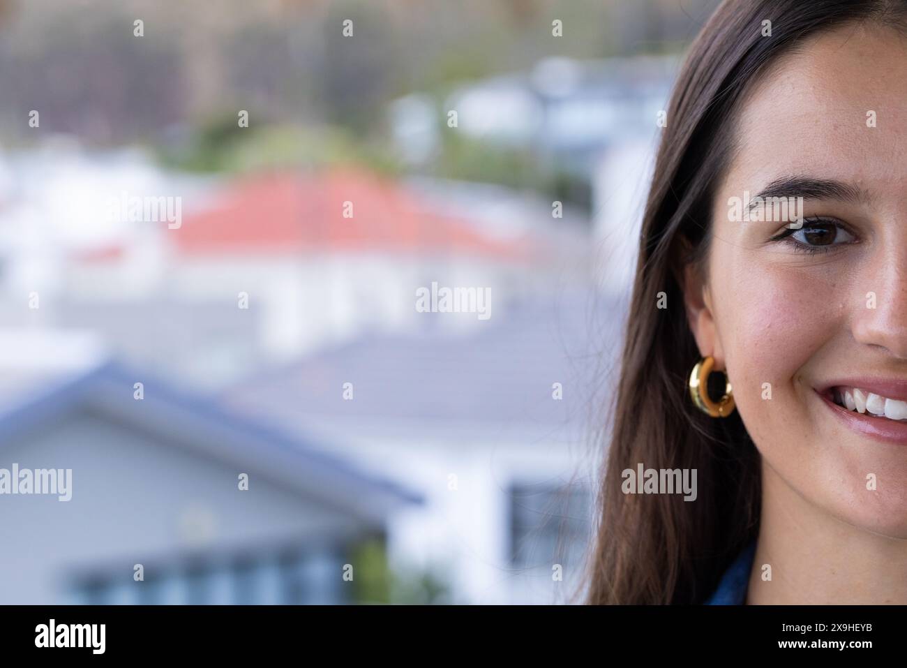 Zu Hause, junge kaukasische Frau mit langen Haaren, die warm lächelt, draußen steht, Kopierraum. Verschwommene Häuser im Hintergrund deuten auf eine Wohngegend hin, cr Stockfoto
