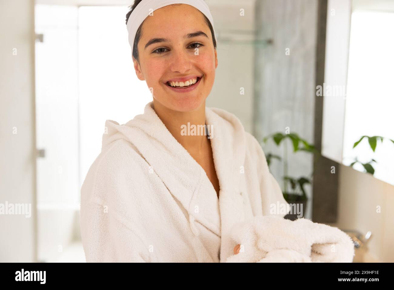 Zu Hause lächelt die junge kaukasische Frau in einem weißen Bademantel und Haarreif. Ein Handtuch in einem hellen Badezimmer mit modernen Armaturen und grünen Pflanzen Stockfoto