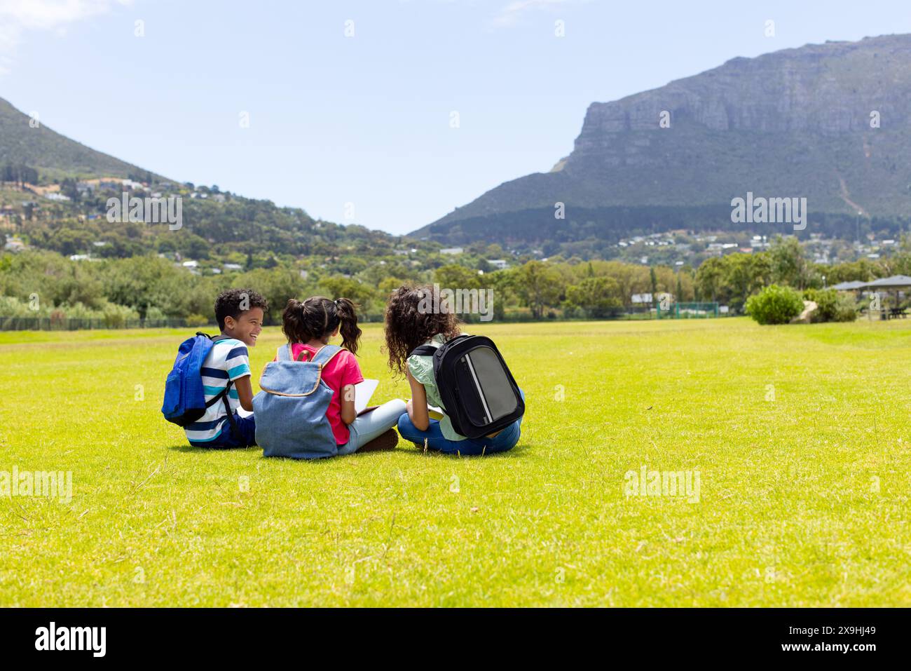 Drei Kinder sitzen auf einem Grasfeld, Rucksäcke auf und nach außen gerichtet, mit Kopierraum Stockfoto