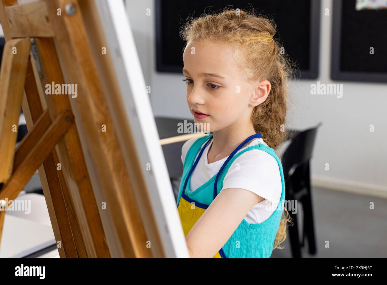 Weißes Mädchen mit lockigen blonden Haaren malt in der Schule auf einer Staffelei Stockfoto