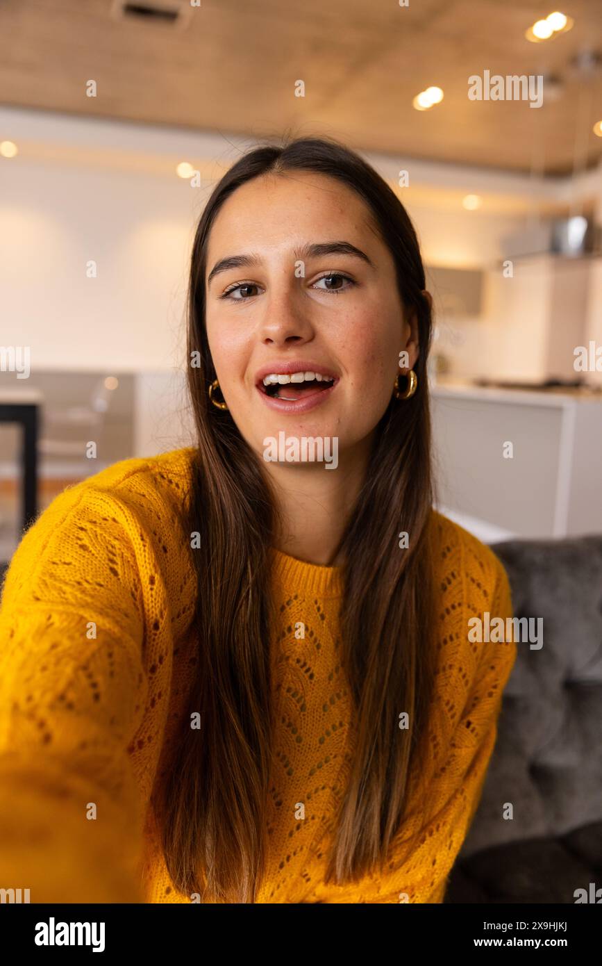 Bei Videoanruf zu Hause lächelt eine junge weiße Frau mit langem, gelbem Pullover Stockfoto