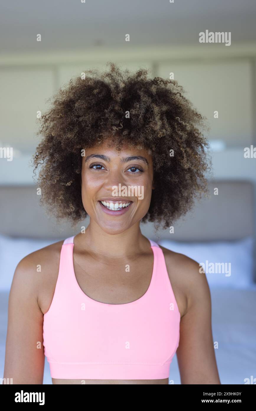 Zu Hause, zweirassige junge Frau, die ein rosa Oberteil trägt, lächelt im Schlafzimmer Stockfoto