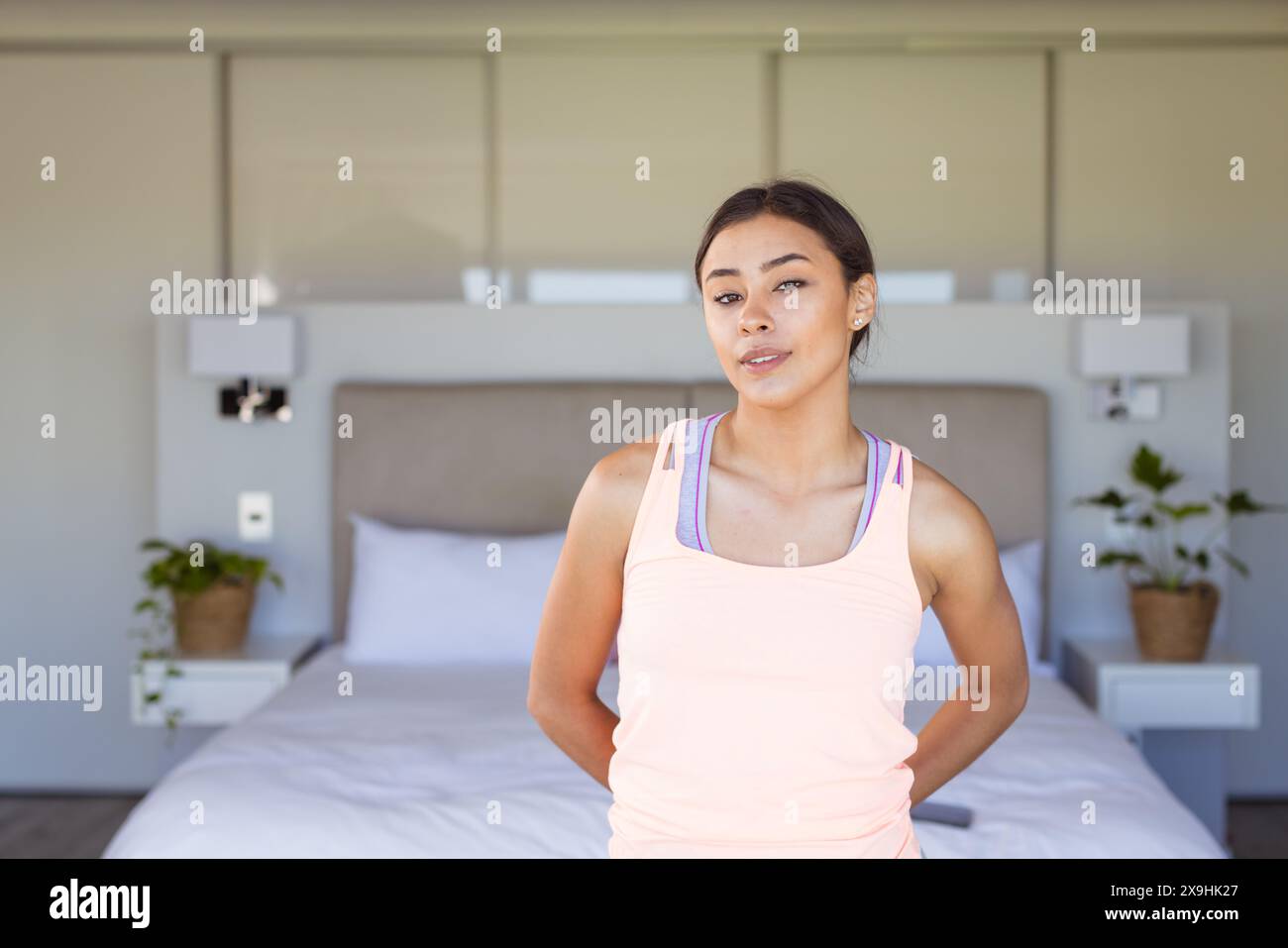 Zu Hause, zweirassige junge Frau mit rosa Tanktop, stehend im Schlafzimmer Stockfoto