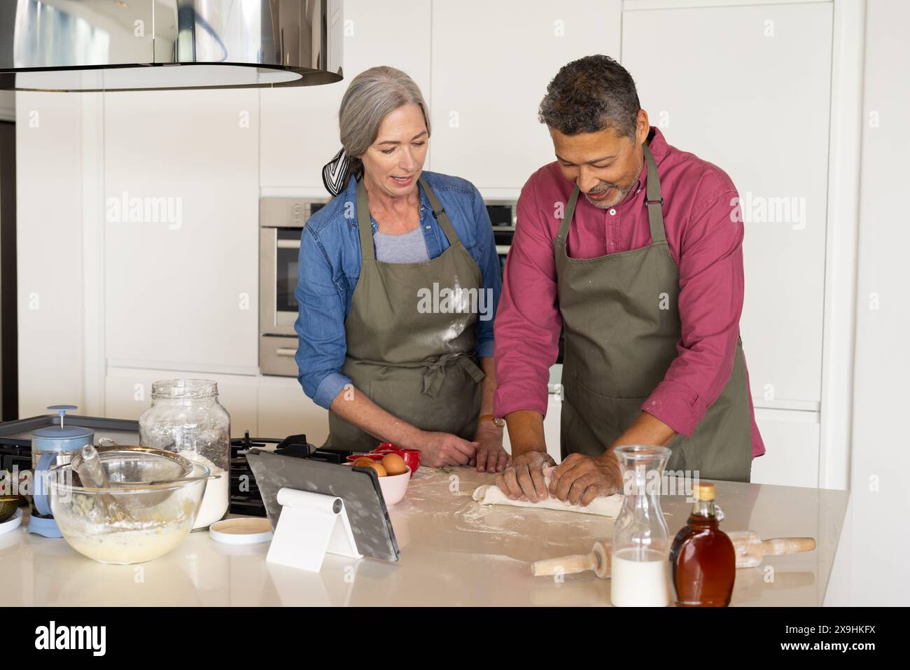 Zu Hause backen verschiedene Seniorenpaare in der modernen Küche zusammen, beide in Schürzen. Kaukasische Frau mit grauem Haar und birassischer Mann mit kurzen dunklen Haaren Stockfoto