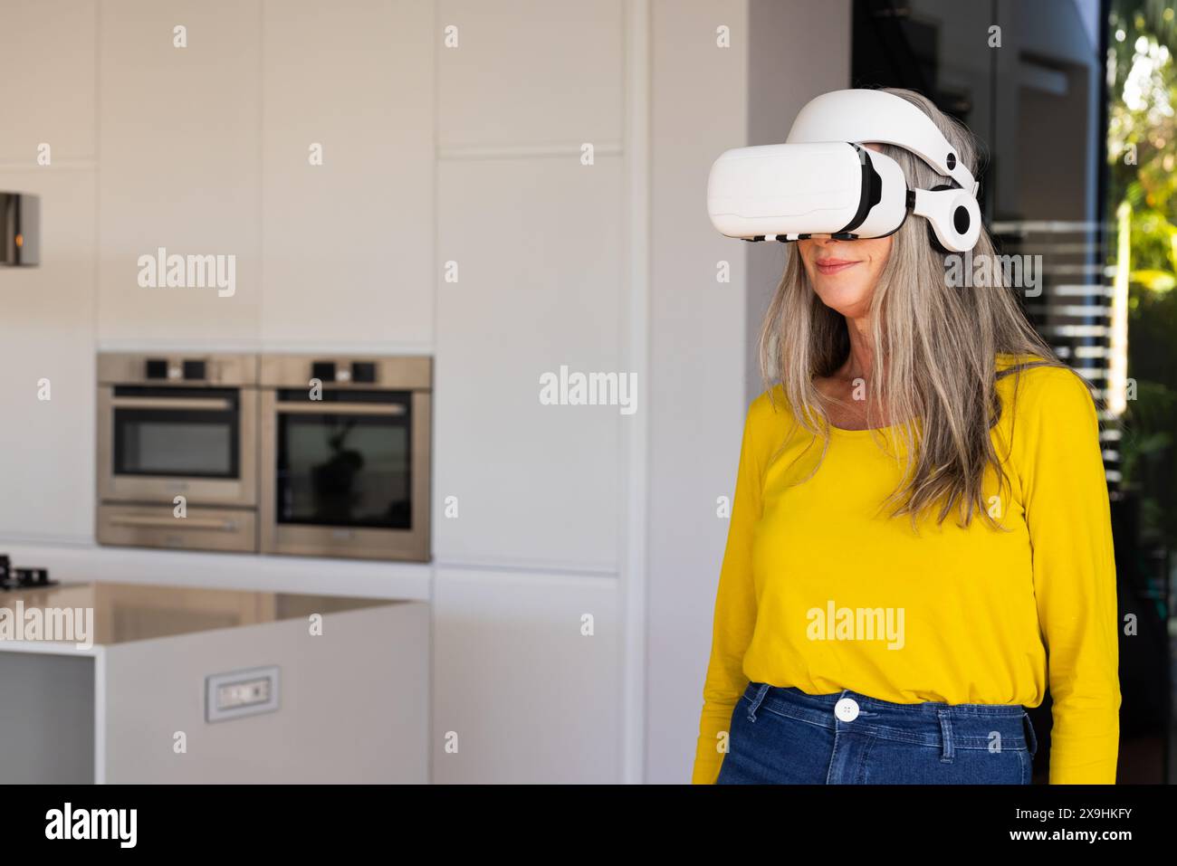 Zu Hause trägt eine kaukasische Seniorin ein Virtual-Reality-Headset in einer modernen Küche, Kopierraum. Lächelnd und mit gelbem Hemd und Jeans Stockfoto