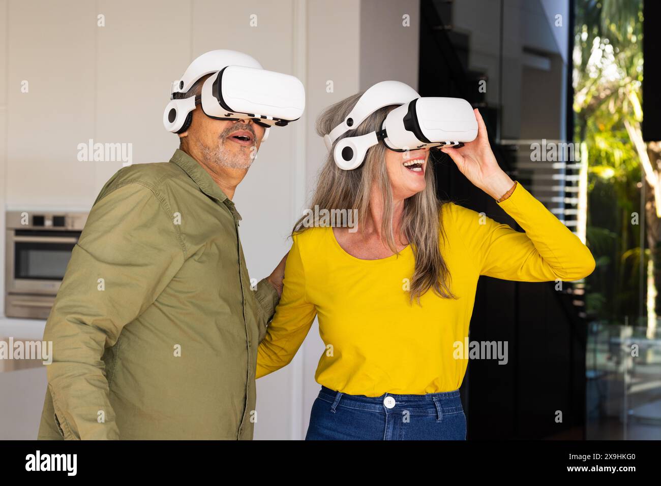 Zu Hause ein vielseitiges Seniorenpaar mit Virtual-Reality-Headsets in der modernen Küche. Lächelnd und umschauend, scheinbar virtuelles Erlebnis zu genießen Stockfoto