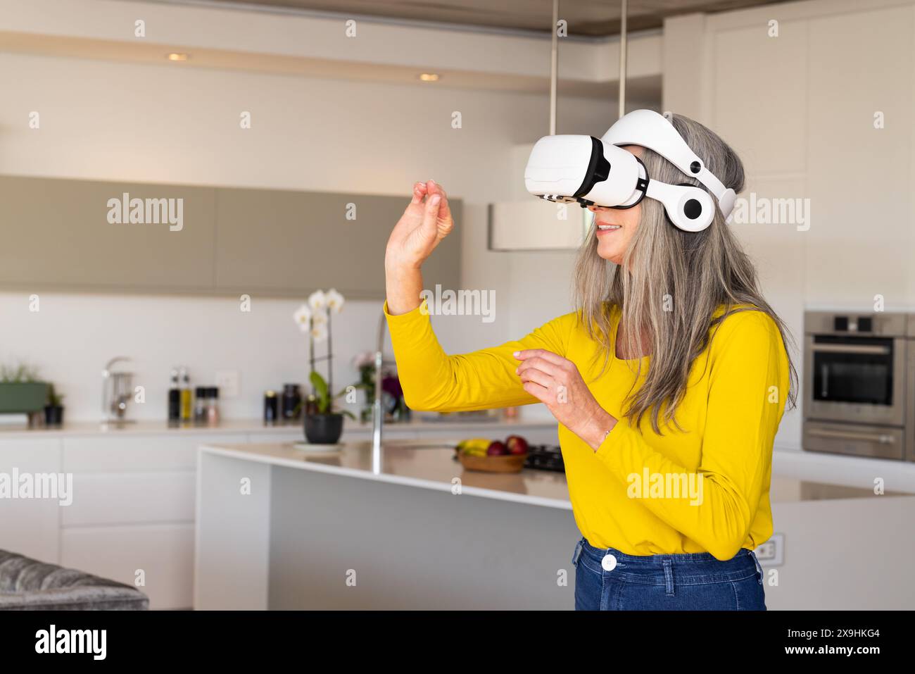 Zu Hause, kaukasische Seniorin in der modernen Küche mit VR-Headset, lächelnd, Kopierraum. Mit gelbem Hemd und Jeans und virtuellem umfeld Stockfoto