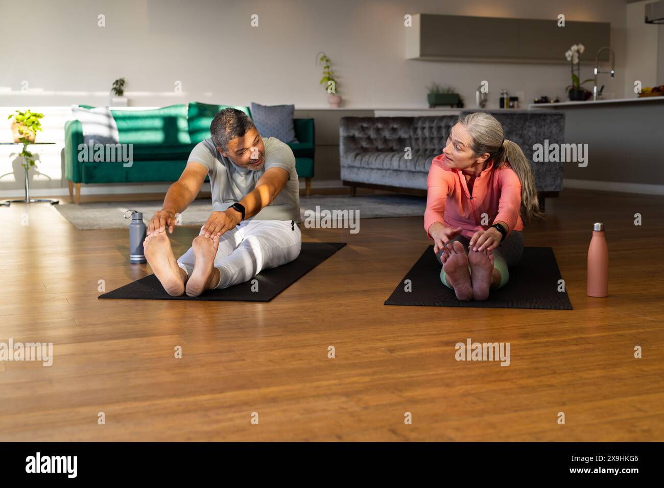 Zu Hause ein vielseitiges Seniorenpaar, das sich auf Yoga-Matten in einem hellen Wohnzimmer ausdehnt. Birassischer Mann mit kurzen dunklen Haaren und kaukasische Frau mit langem grauen hai Stockfoto