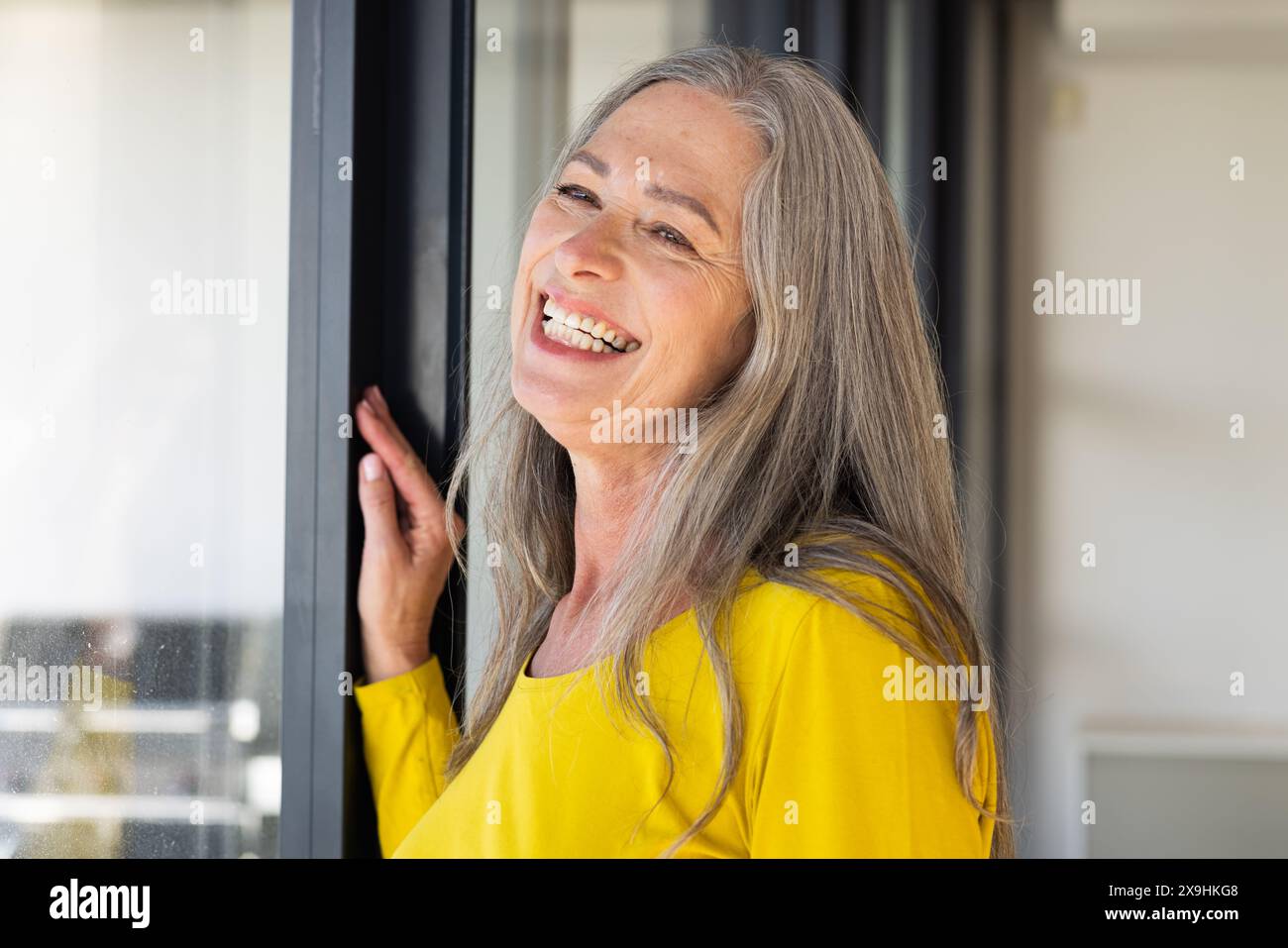 Zu Hause eine kaukasische Seniorin mit langen grauen Haaren und Falten, die am Fenster lächeln. Ein hellgelbes Oberteil, das glücklich und entspannt in einem Heim wirkt Stockfoto