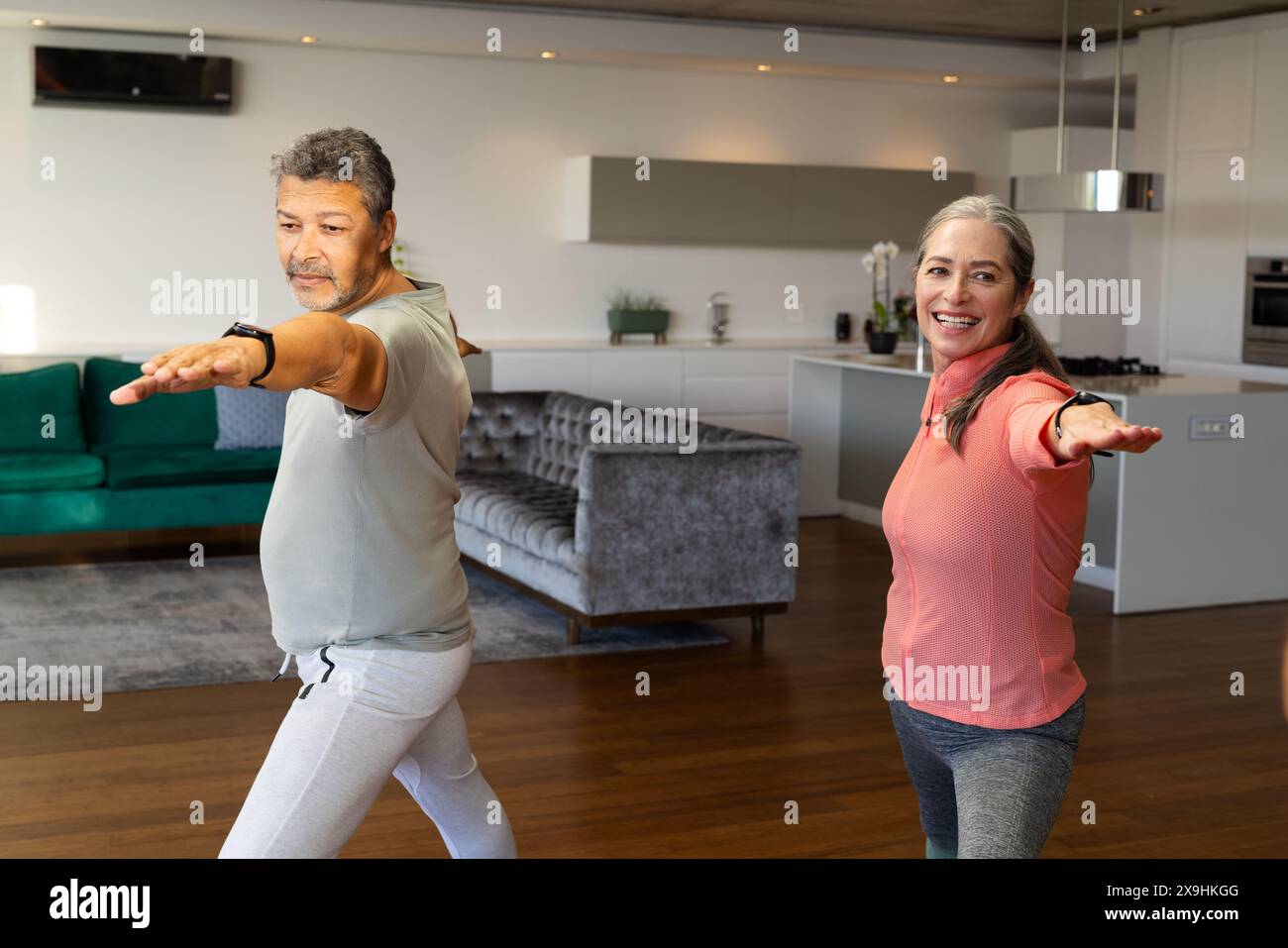 Zu Hause praktiziert ein vielseitiges Seniorenpaar gemeinsam Yoga in einem modernen Wohnzimmer. Reife kaukasische Frau mit langen grauen Haaren und älterer birassischer Mann mit Stockfoto
