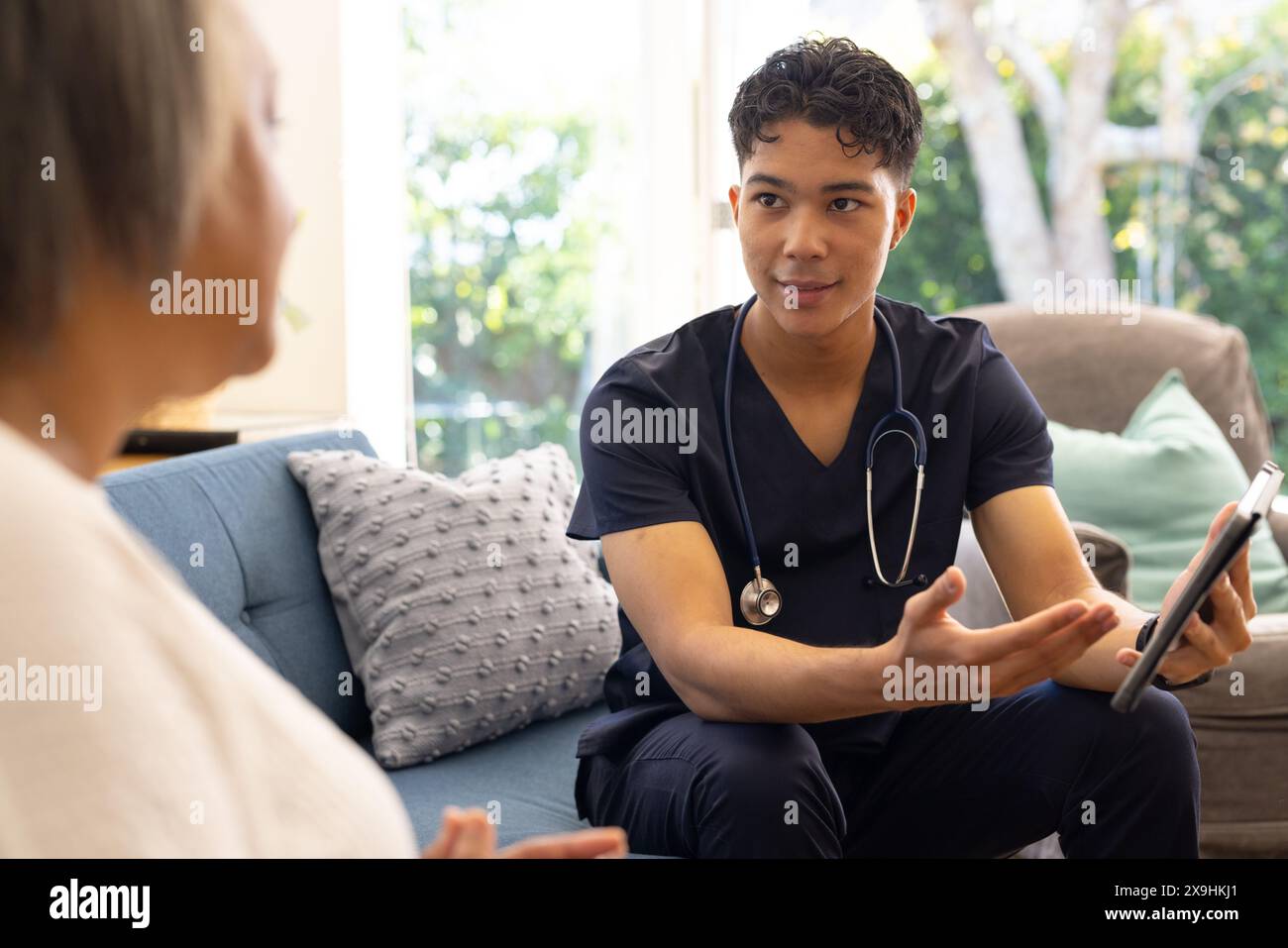 Zu Hause erklärt die birassische männliche Krankenschwester der älteren Patientin aufmerksam. Sie befinden sich in einem gemütlichen Wohnzimmer mit einem großen Fenster, das einen üppigen Garten zeigt Stockfoto