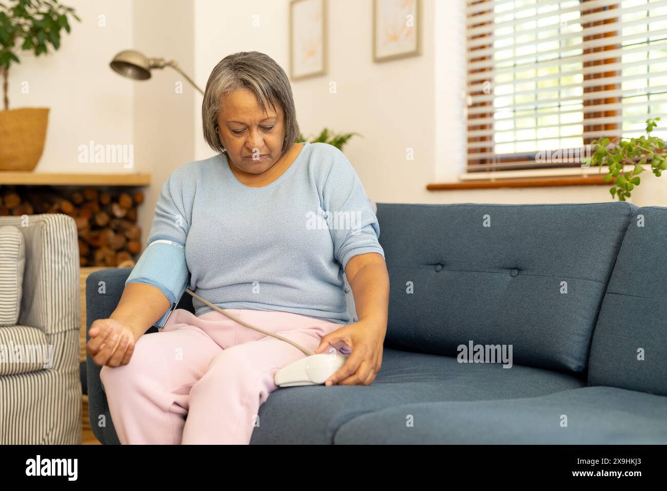 Zu Hause, zweirassige Seniorin, die den Blutdruck auf der Couch im Wohnzimmer überprüft, im Kopierraum. Moderne Einrichtung und Tageslicht, die Raum einfüllt, schaffen eine angenehme Atmosphäre Stockfoto